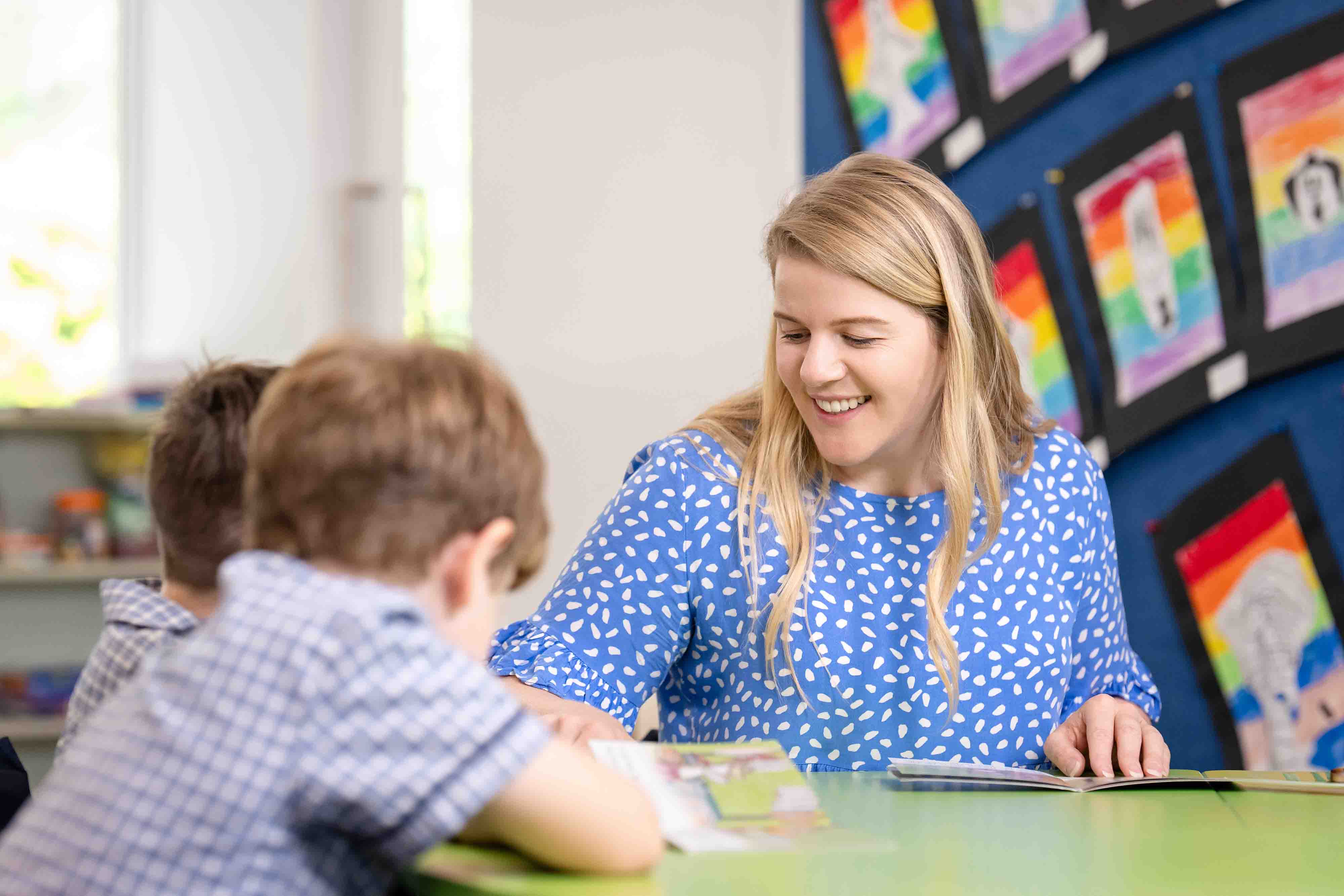 Adult seated at a table reading with students during a classroom learning activity.