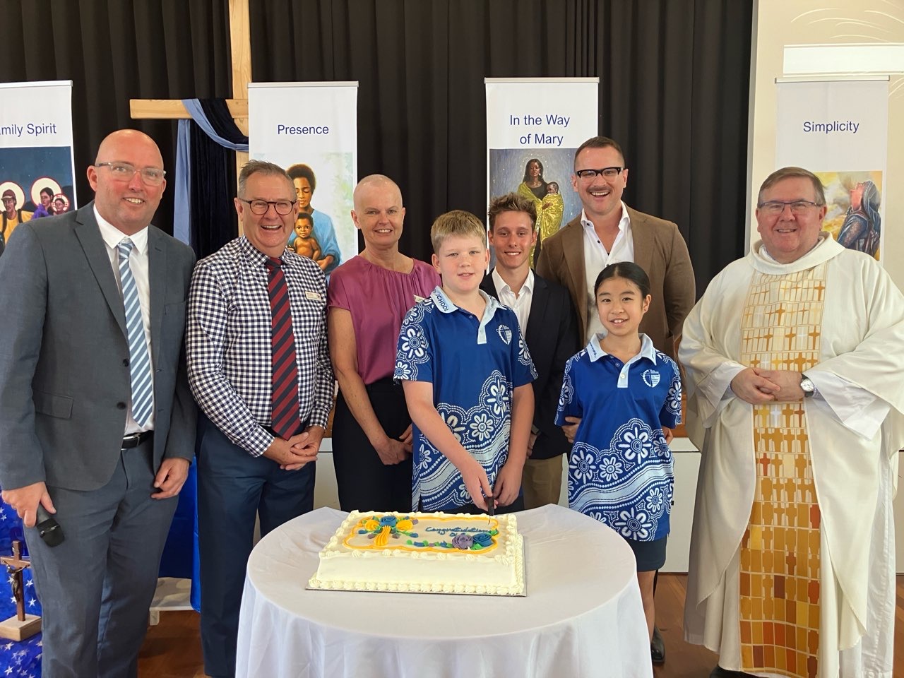 School community group gathered around a celebration cake indoors.