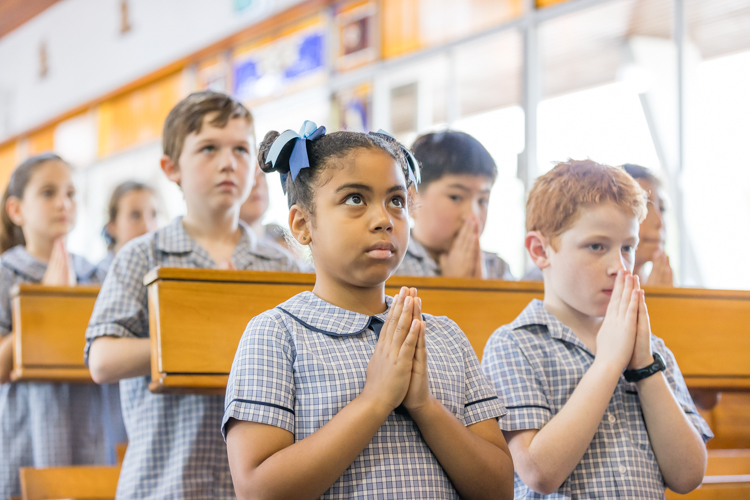 Students in school uniform standing in church pews with hands together during prayer.