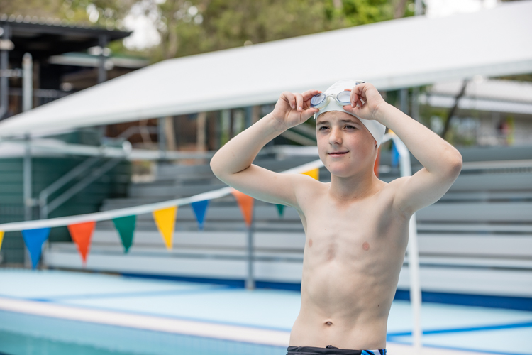 Student adjusting swim goggles beside a school swimming pool.