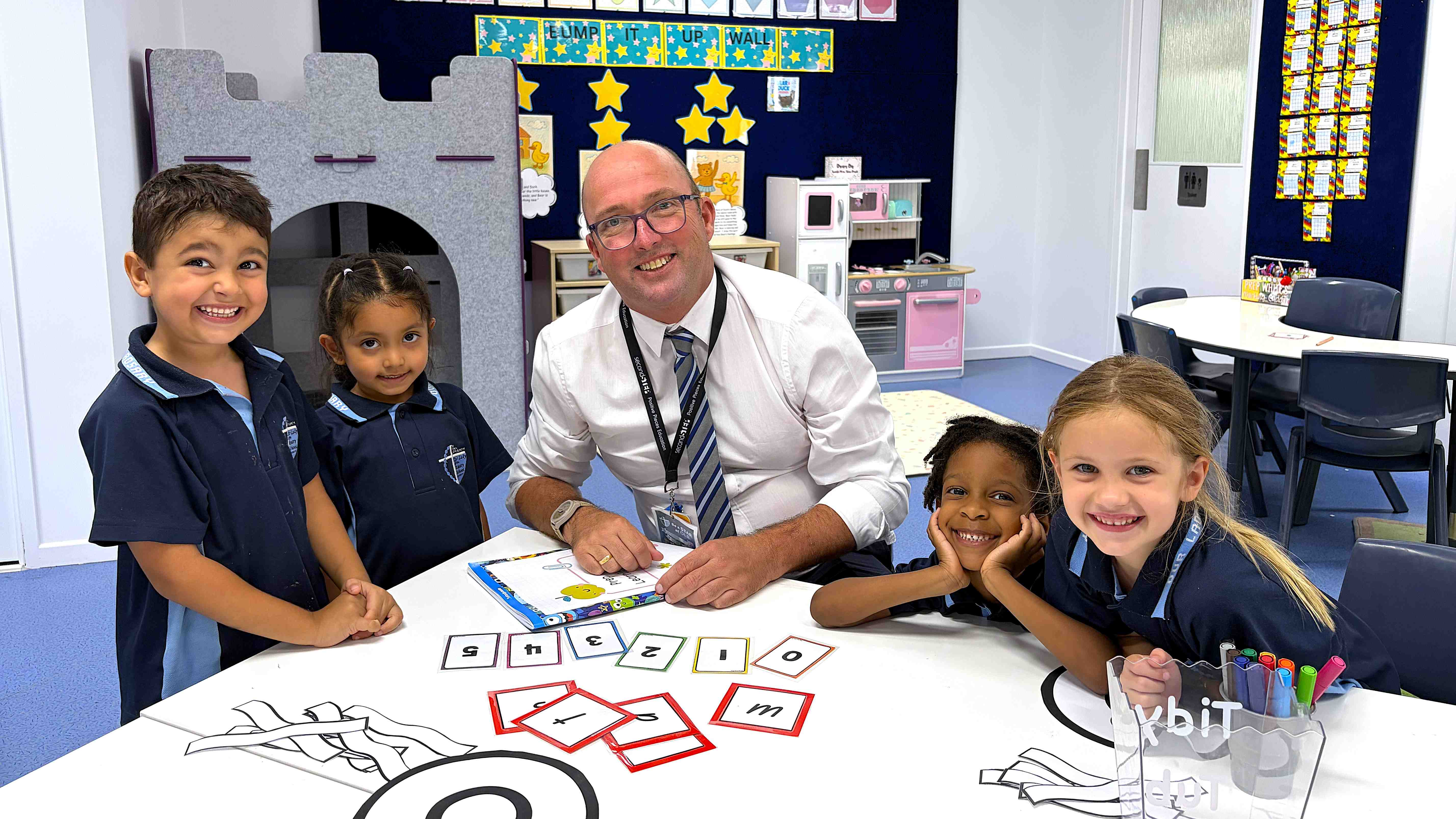 Adult seated at a table with young students working on letter cards during a classroom learning activity.
