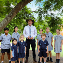 Adult standing outdoors with a group of students in school uniform beneath a large leafy tree.