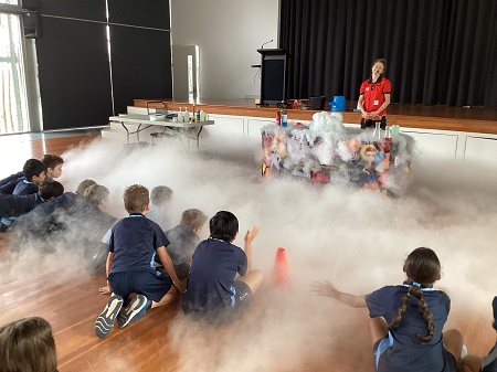 Students seated on the floor watching a science demonstration with fog in a school hall.
